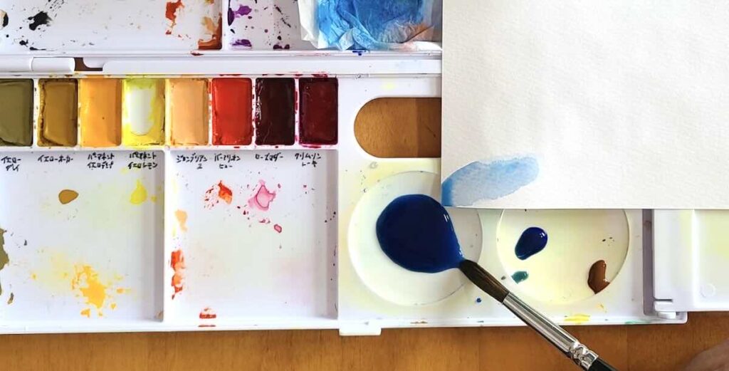 白い水彩パレットの大きな混色スペースに、たっぷりと作られた濃い青色の絵の具。筆の先には絵の具が滴りそうなほど含まれており、隣の水彩紙には鮮やかな青のウォッシュが試し塗りされています。