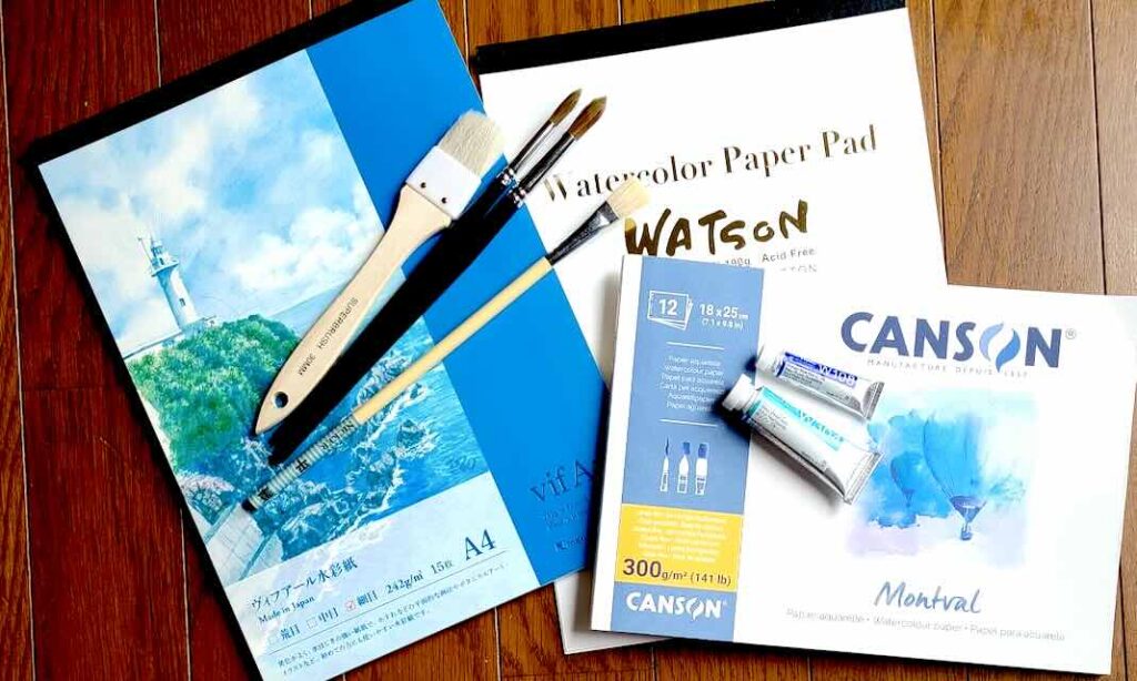 水彩画で空を描くために必要な、平筆、刷毛(ハケ)、丸筆が、ヴィフアールやキャンソンの水彩紙パッドの上に並べられている俯瞰写真。